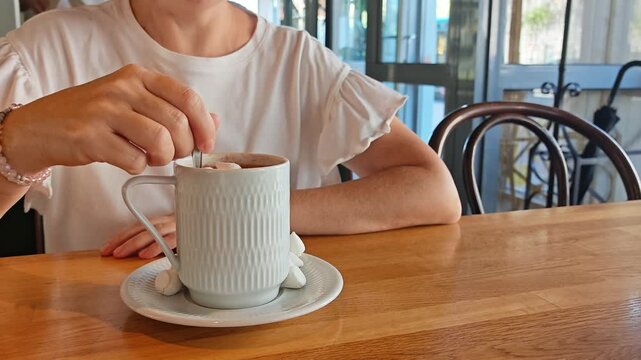 Person enjoying cocoa topped with marshmallows in a white cup on a saucer at a wooden cafe table, cozy winter treat, relaxed lifestyle, sweet comfort, cafe moment.