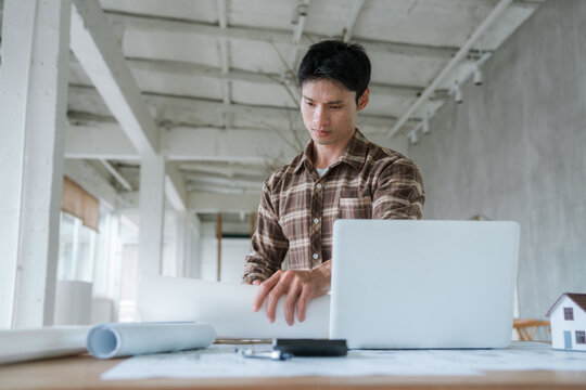 Architect working at a laptop with architectural blueprints in a modern studio, representing building design, construction planning, engineering, and professional project development.