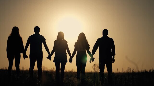 Silhouette group holding hands at sunset. Five people stand together showing unity and solidarity. Friends demonstrate teamwork outdoors. Silhouettes display connection and support at golden hour.
