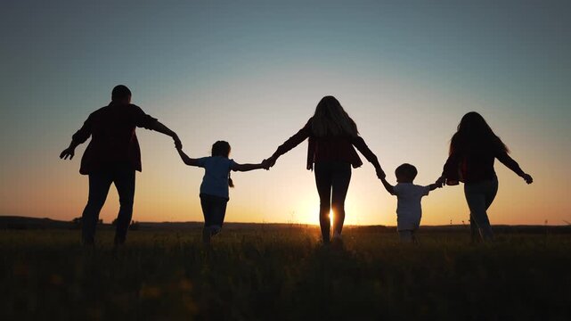 Silhouetted family of five holding hands walking through countryside field at golden hour sunset. Parents and children create connected chain while enjoying peaceful evening nature walk together.