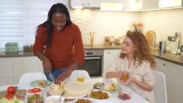 Two happy women preparing and eating burritos in a home kitchen