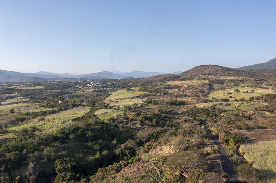 Aerial view of the sugar cane landscape with rolling hills, lush green fields, a winding road, and a distant town in Taretan, Michoacan, Mexico.