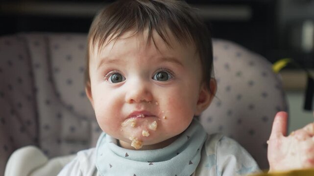 Adorable messy baby with blue eyes eating pureed food in highchair during mealtime. Parent's hand feeds infant with spoon. Toddler has food on face while learning to eat solid foods at home.