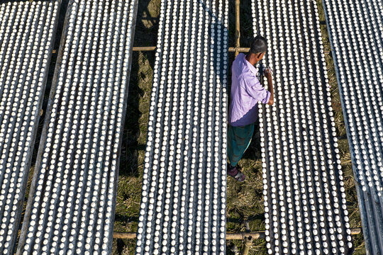 Aerial view of a worker arranging rows of small white food items on bamboo drying trays in Gaibandha, Rangpur Division, Bangladesh.