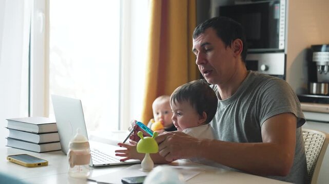 Father multitasking on laptop while holding toddler with baby sitting in highchair at home kitchen table. Dad works typing on computer managing childcare. Parent multitasks with two young children.