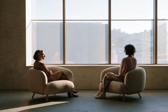 A sunlit office space with two armchairs. A therapist is sitting in one, listening, while the client is looking out the window, processing thoughts.