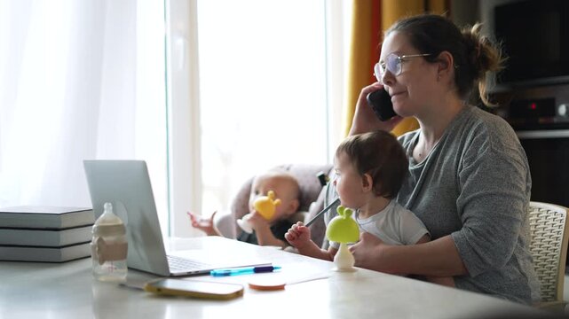 Mother multitasking while working from home with two young children. Woman works on laptop and talks on phone. Mom balances remote work with childcare. Parent manages business tasks with kids.
