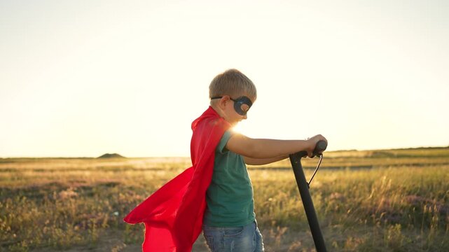 Superhero child rides scooter in open field at sunset. Boy in red cape and mask plays hero outdoors. Superhero kid grips scooter handle as cape flows. Child hero plays at golden sunset.