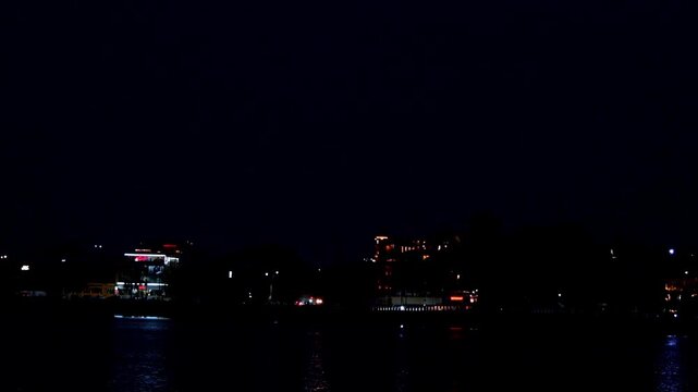 Distant building lights scatter soft reflections on the lake. Dark skies hover over the quiet water. Peaceful night scenes cover Udaipur, Rajasthan.