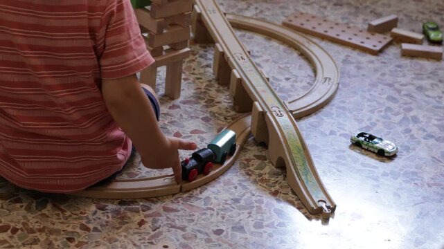 Toddler boy sitting on the floor and pushing a wooden toy train on a railway track, shot from behind. Fine motor skills, montessori learning, and creative indoor play concept
