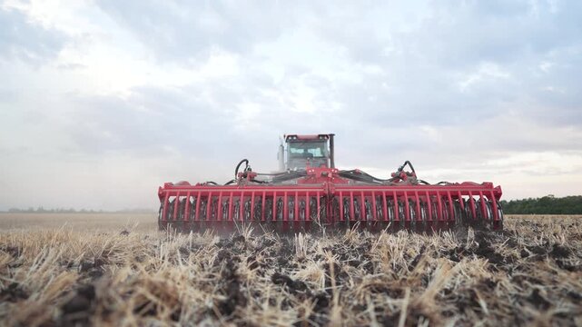 Red tractor with disc harrow cultivating stubble field. Agricultural machinery working on farmland. Tractor tilling soil after harvest. Farming equipment preparing ground. Field cultivation.