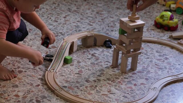 Cute toddler boy playing with a wooden train set on the floor at home while father's hand builds a wooden block tower. Family interaction and creative play concept