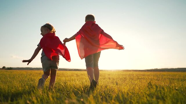 Children playing superhero running through golden field at sunset wearing red capes. Kids hold hands pretending heroes outdoors. Children play with dog in meadow. Superhero game at sunset.