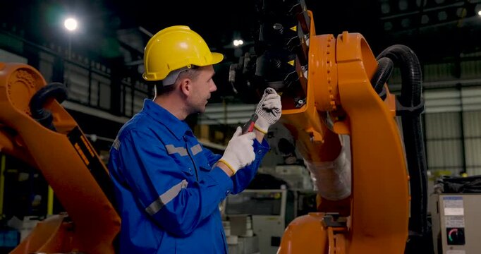 Adult male technician inspects industrial robotic arm with handheld tool in factory workshop, checking mechanical connection and joint condition during preventive automation maintenance service.