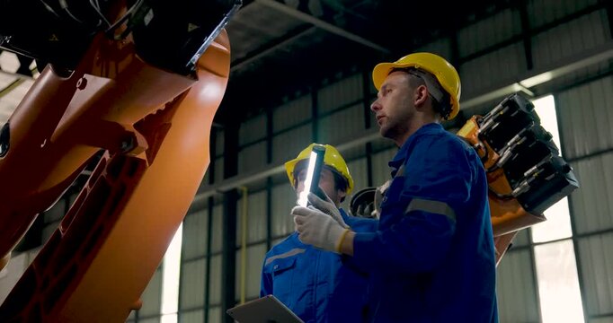Two adult male technicians inspect industrial robotic arm in factory, shining flashlight and pointing at mechanical components while checking machinery condition during preventive maintenance service.