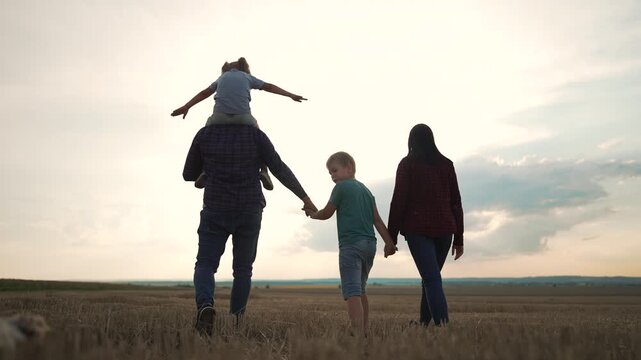 Family walking together in field at sunset with child on shoulders holding hands outdoors. Parents carry daughter while son walks between them. Family enjoys nature walk at dusk in countryside.