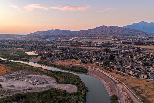 Aerial view of the Jordan River Parkway winding through residential neighborhoods and Silicon Slopes at sunset with distant mountains under a soft sky in Lehi, Utah, United States.