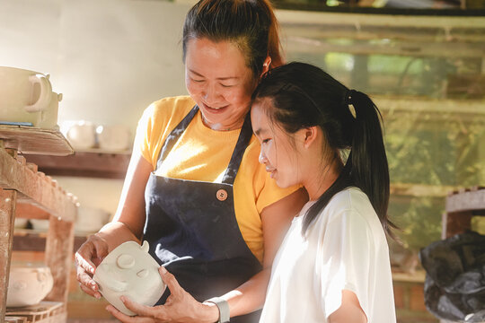 Smiling Asian mother showing handmade smiling face ceramic cup to daughter in pottery studio, sweet mom teaching kid craft, loving parent child bonding, family small business workshop lifestyle