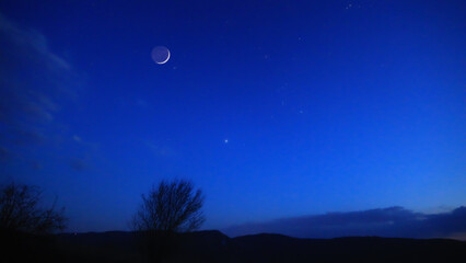 Blue hour time with crescent Moon, stars and planets above landscape silhouettes. © astrosystem