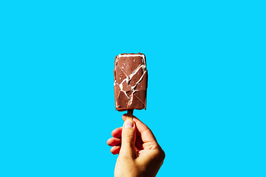 Hand Holding Chocolate Ice Cream Bar with Cracked Shell on Blue Background