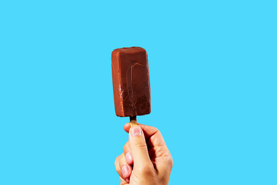 Hand holding chocolate ice cream bar on wooden stick against blue background