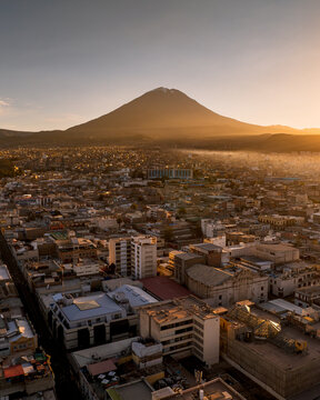 Aerial view of the city of Arequipa at sunset with the Misti volcano in the background, featuring urban rooftops, historic buildings, and soft golden light in Arequipa, Arequipa, Peru.