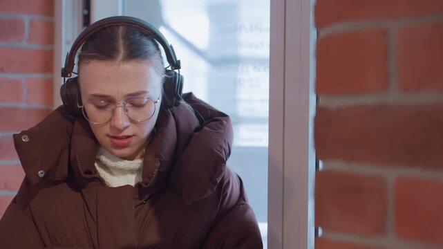 White student listening in cafe corner with headphones and glasses, seated against exposed brick near window, thoughtful expression and calm posture convey concentration and quiet reflection, low