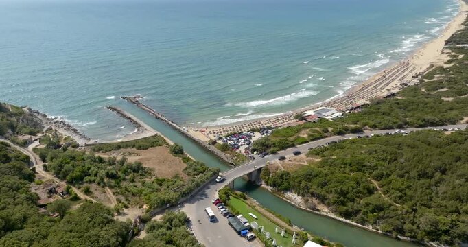 Aerial view of the coastal canal entrance in Sabaudia, Lazio, Italy, with a bridge crossing the water toward turquoise waves crashing on a sandy beach surrounded by lush green landscape.