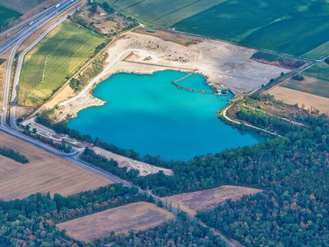 Aerial view of a turquoise quarry lake surrounded by industrial excavation sites, green fields, and a nearby highway in Sausheim, Grand Est, France.