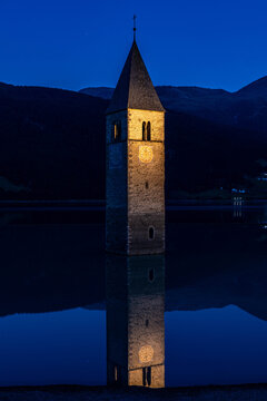 Illuminated sunken bell tower of Curon reflecting in the calm waters of Lake Resia at dawn, South Tyrol, Italy