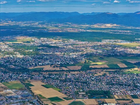 Aerial view of the town of Sausheim with agricultural fields, forests, and distant mountains under a blue sky in Sausheim, Grand Est, France.