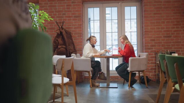 Cozy cafe Black woman White woman sharing animated conversation at brick wall window, sipping coffee and gesturing, laughing over latte on small table, plants and modern chairs, warm daylight