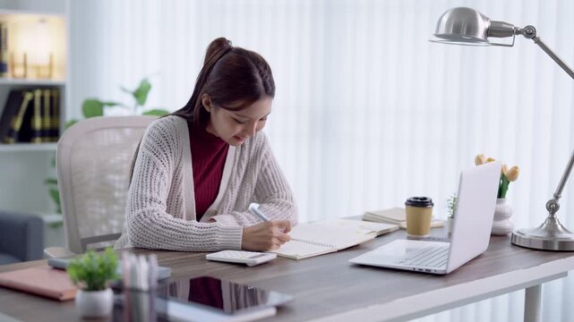 Young Asian woman writing notes in notebook while studying with laptop at home office desk