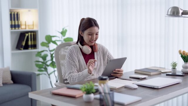 Young Asian woman in headphones using tablet for online learning at bright modern home desk