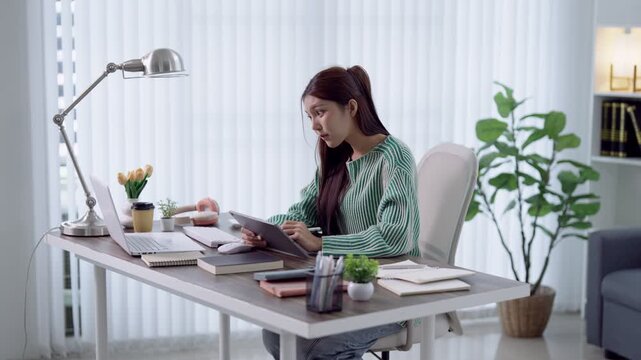 Young Asian woman studying with digital tablet at modern home office desk surrounded by books and plants