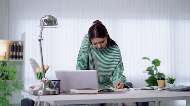 Young Asian student writing notes while studying with laptop at bright home office desk