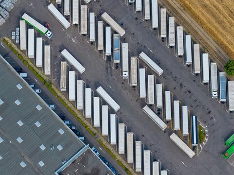 Aerial view of a logistics center in Sausheim, featuring rows of parked semi-trailer trucks and trailers in a paved depot area next to a warehouse. Sausheim, Grand Est, France.