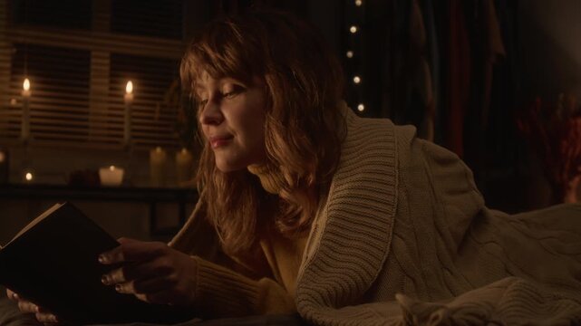 Young Caucasian woman lying on bed and reading book during lightning flash flickering outside window. Footage ideal for promoting mental health, reading habits, or wellness brands