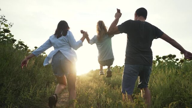 Happy family running through sunflower field at sunset. Parents hold daughter hands and lift child in air. Family runs along dirt path together. Happy parent and kid enjoy summer walk outdoors.
