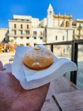 Tette delle Monache cream-filled pastry dusted with powdered sugar held in hand on napkin, Bari old town square and church tower visible in background