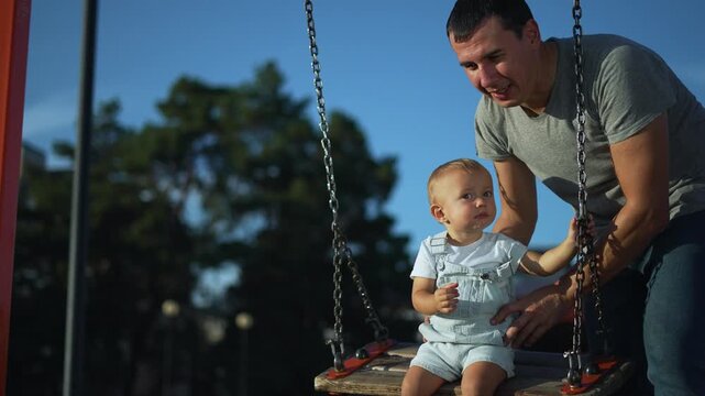 Smiling father in gray t-shirt gently pushes curious blond baby wearing overalls on a chain swing at an outdoor playground. Dad and toddler enjoy bonding time under a clear blue evening sky.