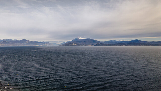 Aerial view of Lake Garda with snow-capped mountains in the background under an overcast sky in Sirmione, Lombardy, Italy.