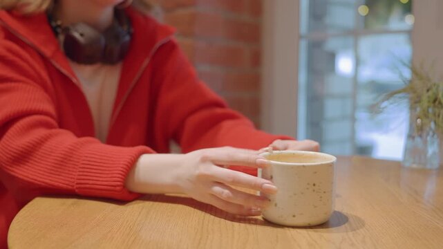 Woman holding ceramic mug at cafe table student pausing between study sessions, writer sketching ideas, remote worker listening to music red sweater, headphones around neck, natural window light,