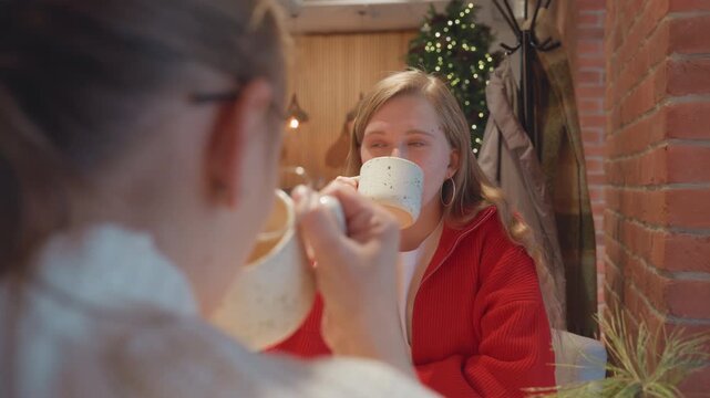 Young white woman in cozy cafe sipping speckled mug across friend, warm brick wall backdrop, soft string lights and greenery, relaxed smile during friendly conversation, eye contact and candid