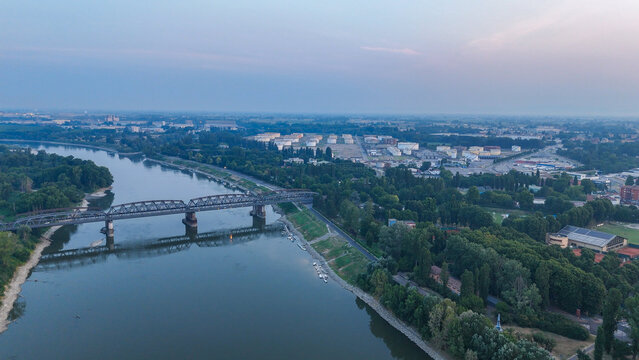 Aerial view of the Po River in Cremona during a summer heatwave with low water levels, a historic iron railway bridge, and industrial storage tanks in the background, Cremona, Lombardy, Italy.