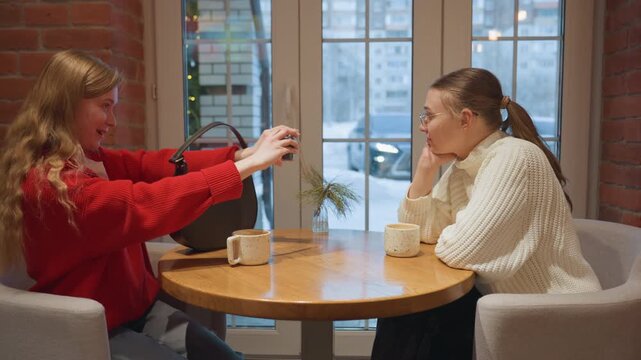 Two white friends at cafe table exchanging phone and laughing, red sweater and cream sweater contrast, mugs on wooden table, brick wall and city window backdrop, hands meet during photo share, warm