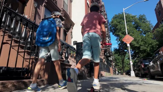 Two boys, one walking with a backpack and one on a scooter, returning from school along a typical brownstone-lined sidewalk on a sunny day in brooklyn, new york city