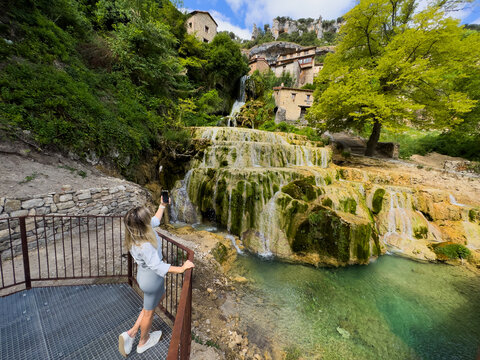 woman taking photograph of cascading waterfall in serene natural landscape behind stone steps and railing with peaceful and calm atmosphere and vibrant green trees and brown stone. Cascada de Orbaneja