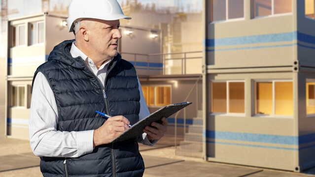 Modular Construction. Site engineer inspects modular building units and records project progress on a clipboard during residential development supervision.