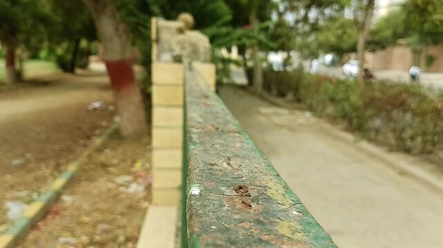 Macro perspective shot along a rustic green painted metal fence rail in a public park with heavily blurred trees out back